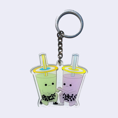 Acrylic die cut keychain of an illustrated pair of boba cups with arms and legs. They both are down on one knee and clasp a hand together. One is green and the other is purple.