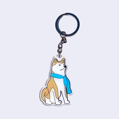 Die cut clear acrylic keychain of an illustrated brown and white Shiba Inu dog, sitting with a blue scarf around its neck.