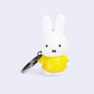 Figure of Miffy, standing with her arms to her side and her classic expression of open eyes and an "x" mouth. She is white and wears a yellow dress.