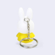 Figure of Miffy, standing with her arms to her side and her classic expression of open eyes and an "x" mouth. She is white and wears a yellow dress.