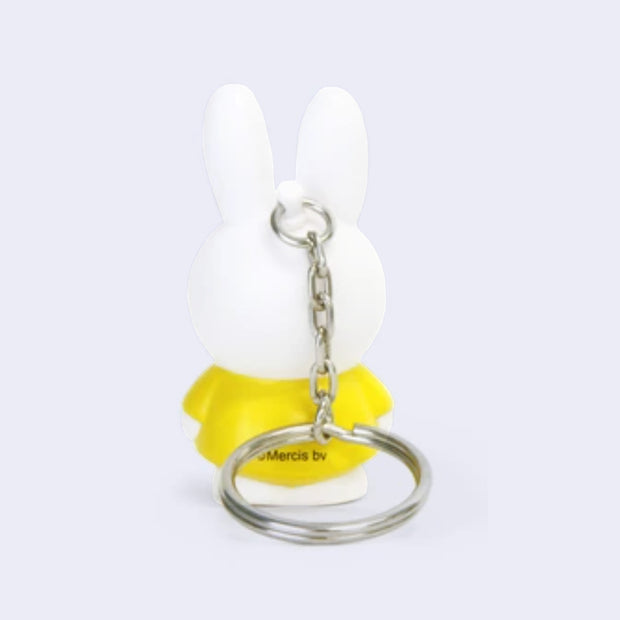 Figure of Miffy, standing with her arms to her side and her classic expression of open eyes and an "x" mouth. She is white and wears a yellow dress.