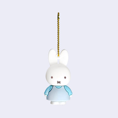 Rubber mascot figure of Miffy, wearing a light blue dress with a blue long sleeve underneath. The figure is attached to a gold toned ball chain.