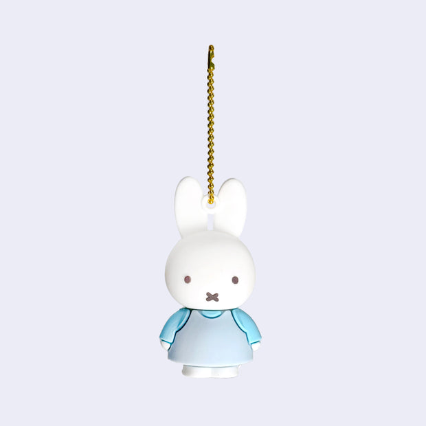 Rubber mascot figure of Miffy, wearing a light blue dress with a blue long sleeve underneath. The figure is attached to a gold toned ball chain.