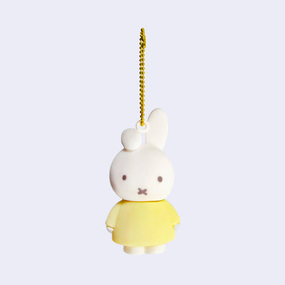 Rubber mascot figure of Miffy, wearing a light yellow dress with one of her bunny ears folded down. The figure is attached to a gold toned ball chain.