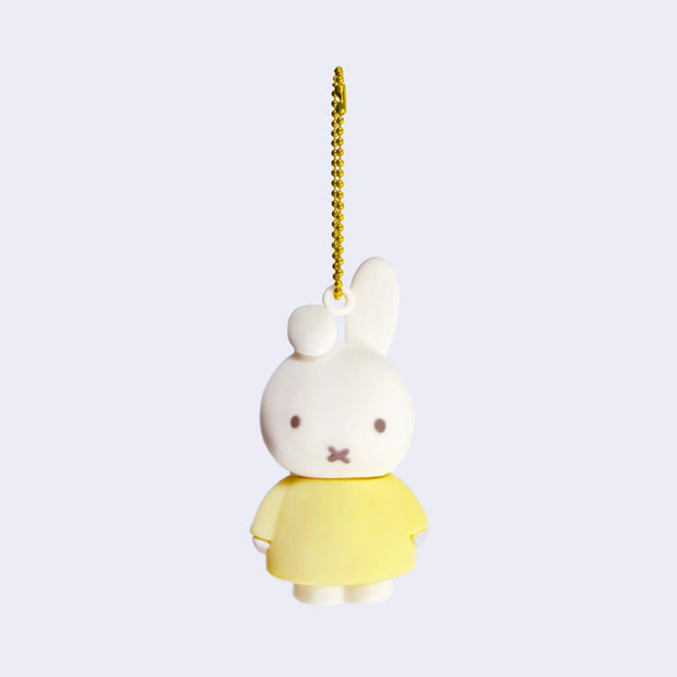 Rubber mascot figure of Miffy, wearing a light yellow dress with one of her bunny ears folded down. The figure is attached to a gold toned ball chain.