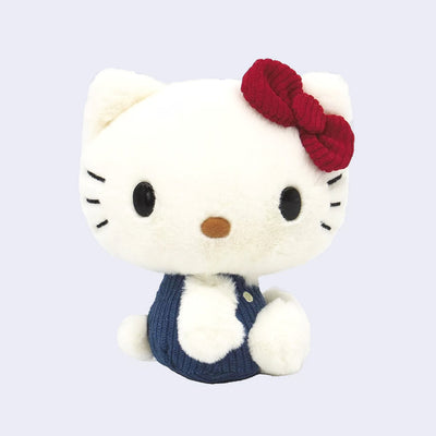 Fluffy white plush doll of Hello Kitty, sitting with her head turned to the side, so her face and body face different directions. She has a burgundy red corduroy hair bow and blue corduroy overalls.