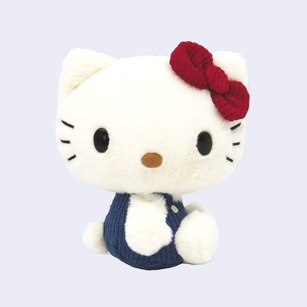 Fluffy white plush doll of Hello Kitty, sitting with her head turned to the side, so her face and body face different directions. She has a burgundy red corduroy hair bow and blue corduroy overalls.