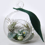 A small, round glass terrarium sits against white background. There is a single felt leaf that hangs from the hanging ring at the top of the terrarium. There is an opening in the glass in the front. Inside there are two air plants, a totoro figuring smiling widely and glass rock and moss.