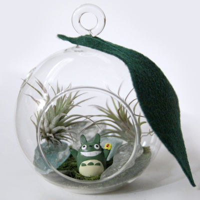 A small, round glass terrarium sits against white background. There is a single felt leaf that hangs from the hanging ring at the top of the terrarium. There is an opening in the glass in the front. Inside there are two air plants, a totoro figuring smiling widely and glass rock and moss.