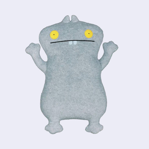 Fluffy gray plush doll of a mostly flat standing monster with yellow eyes and a straight lined, wide mouth with 2 small teeth showing.